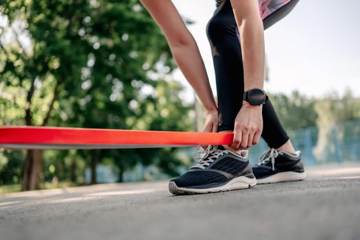 Young girl woman exercising outdoors with rubber elastic band doing training for her legs wearing black leggings and fitness tracker. Active workout with additional sport equipment outside