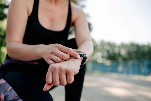 Young girl woman wearing sportwear sitting at stadium and looking at fitness tracker on her hand during exercising. Female person control sport progress with smart watch