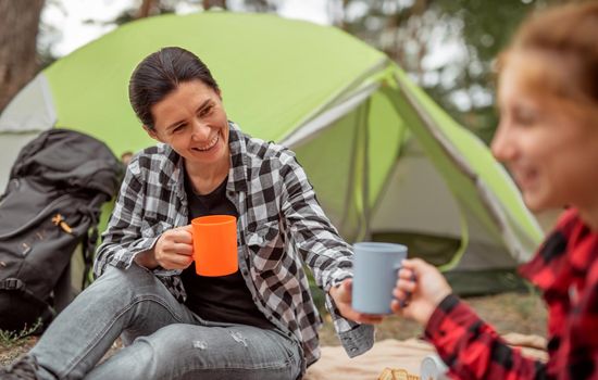 Beautiful woman with girl drinking tea in camping in forest with tent on background and smiling. Young mother and her teenager daughter traveller during hiking in mountains resting