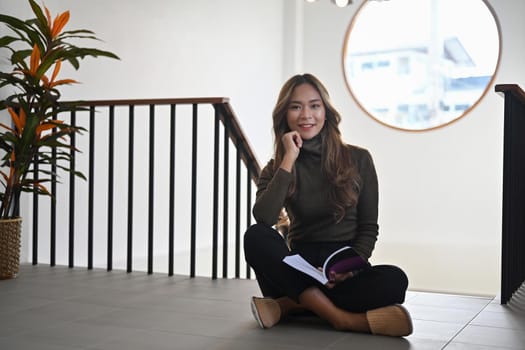 Pleased beautiful asian woman sitting in living room and smiling to camera.