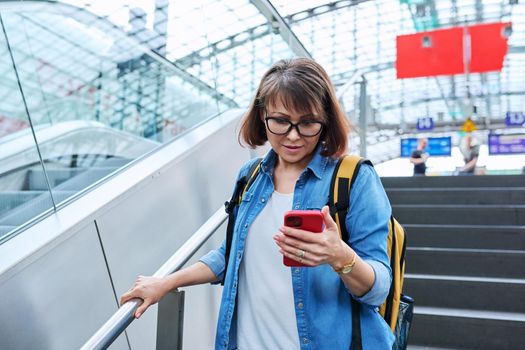 Middle-aged woman with backpack smartphone in her hands walking up stairs, near escalator in modern station building. Urban architecture, urban lifestyle, people concept