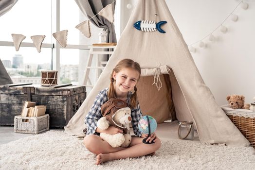 Little girl with teddy bear and globe sitting in wigwam settled in playroom