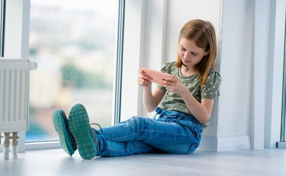 Preteen girl sitting on windowsill with smartphone and looking at display. Beautiful female kid schoolgirl reading from cell phone at home