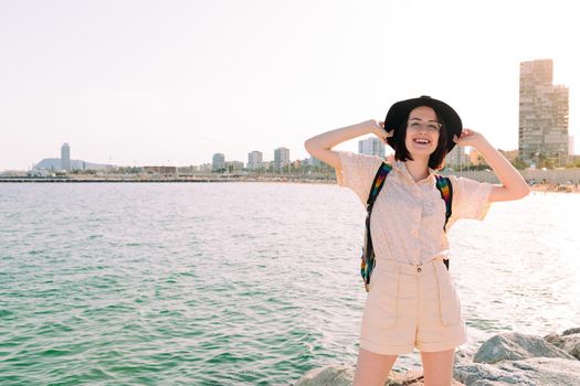 young traveler woman with hat smiles happy near the sea next to the city, travel and tourism concept, copy space for text