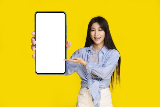 Beautiful asian girl holding smartphone with white screen happy to introduce new app, game, win, isolated on yellow background. Product placement. Mobile app advertisement.