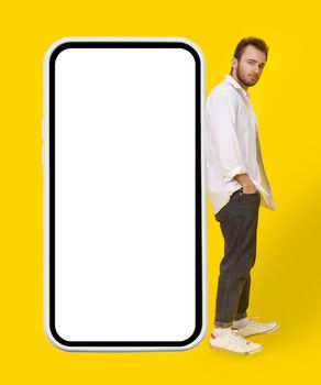 Young man leaned on big smartphone in casual wear and eye glasses posing on yellow background. Stylish bearded smart casual look hipster man with phone. Mobile app advertisement mock up.