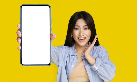 Beautiful asian girl wowing facial expression holding smartphone with white screen happy to introduce new app, game, win, isolated on yellow background. Product placement. Mobile app advertisement.