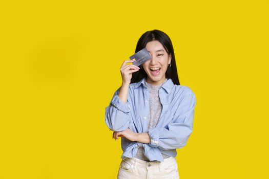 Excited asian girl smiling cover her with bank card right eye ready to spend money. Asian girl in blue shirt with mockup bank card isolated on yellow background. Shopping concept.