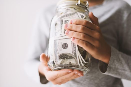 Frugal caucasian teenager boy is holding glass jar piggy bank with money dollars in his hands. Kid save money Saving money for education. Teaching child financial literacy with money Childhood