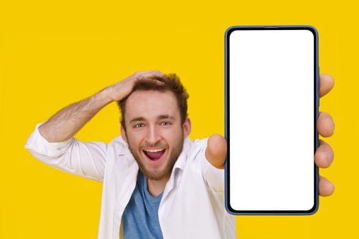 Handsome excited young man emotionally smiling touching his head showing huge smartphone with white screen. Mobile app advertising mockup product placement. Young man with phone.