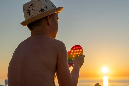 a child plays pop it sitting on the beach in the setting sun. A child playing with a rainbow pop it, presses the bubbles with his fingers. anti-stress toy for children and adults