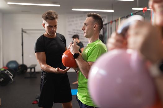 A trainer assists and explains the proper exercise to a young man doing sports and performing squats with the kettlebell in his hands, at the local training and fitness center