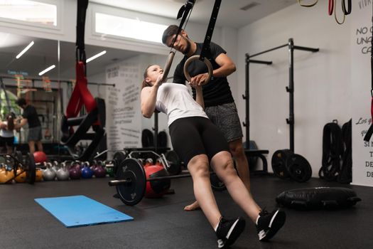 Young woman plays sports and performs single arm ring row and ring hold, at local training and fitness center, together with her personal trainer