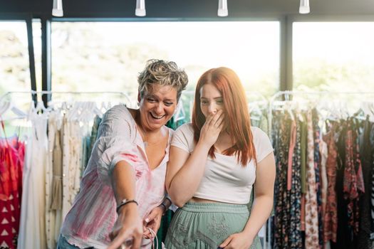 two friends surprised and excited, by exclusive item of clothing in front of them, shopping in a small clothing shop. mother and daughter spending time shopping. shopping concept. natural light from the window of the window.