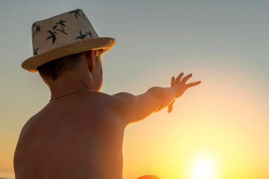 a happy boy in a sun hat sits on the beach at sunset and stretches his hand for the sun. Childhood dreams, family happiness, vacation at sea
