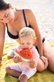 Mom sits on a blanket next to a little girl eating watermelon. High quality photo