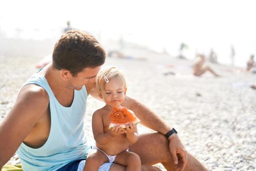 Little girl eating watermelon while sitting on dad lap on the beach. High quality photo