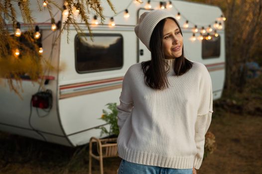 A pensive Caucasian woman in a white knitted sweater and a hat is resting near a motorhome in a warm autumn. Travel by camper