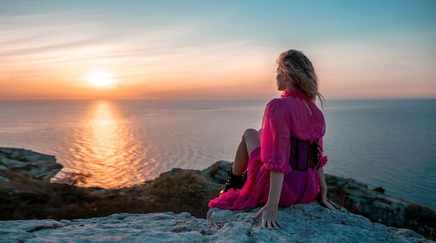 Side view a Young beautiful sensual woman in a red long dress posing on a volcanic rock high above the sea during sunset. Girl on the nature on blue sky background. Fashion photo