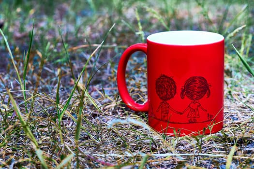 a small bowl-shaped container for drinking from, typically having a handle. Red ceramic cup with inscription love and funny couple on green grass in the park