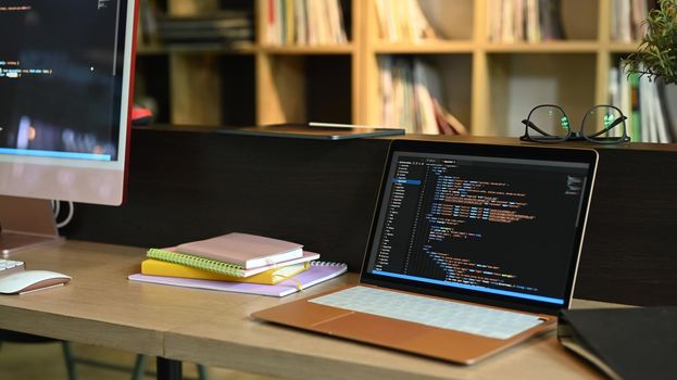 Laptop and computer pc on wooden office desk with programming code on screens.