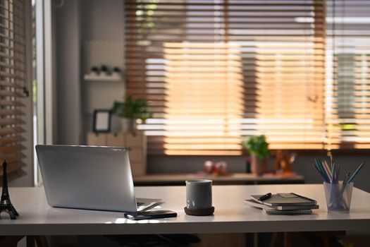 Contemporary home office. Computer laptop, coffee cup, notebooks and pencil holder on white table.