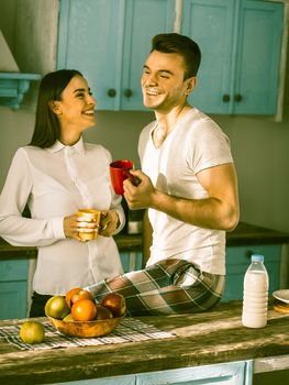 Young Couple Toothy Smiling At Kitchen In Sunny Morning, Happy Man And Woman Laugh Holding Cups With Hot Coffee Or Tea, Woman In Formalwear Looks To Man In Pyjamas Sitting On Table, Toned Image