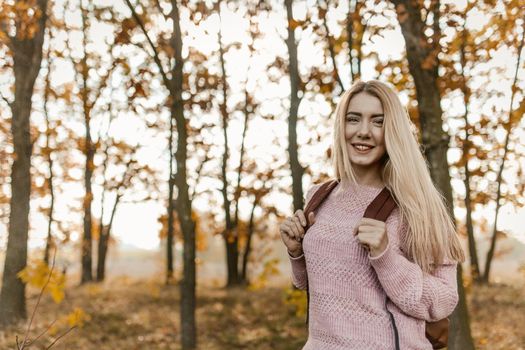 Young Female Traveler Posing On Background Of Autumn Forest Outdoors, Charming Blonde With Backpack Stopped Looking At Camera, Autumn Trees With Yellow Leaves On Sunny Blurry Background