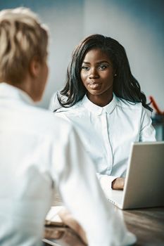 Two Business Women Talking Each Other, Caucasian And African Ladies Talks Sitting At Table, Focus On Face Of Charming African Woman Chatting With Blonde Sitting Back In Foreground, Toned Image