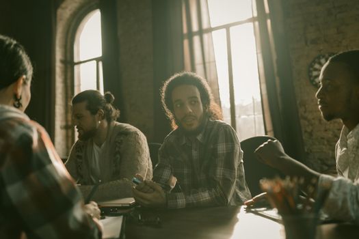 Team Of Young Freelancers Working Together In Workspace, Focus On Arab Guy Sitting In Back Sunlight, Diverse Team Communicates At Table In Stylish Interior With High Arched Windows