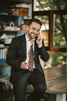 Cheerful Businessman Talks Over Phone Sitting On Desk, Portrait Of Well-Dressed Handsome Macho Talking Over Smart Phone During Coffee Break While Sitting On Table In Modern Office
