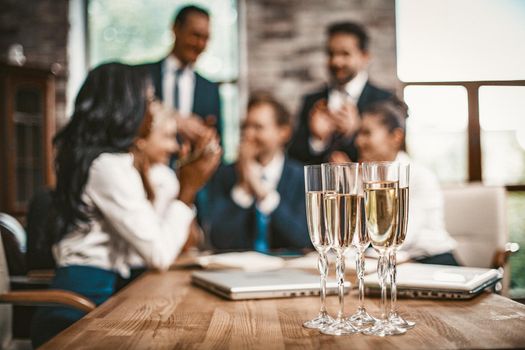 Celebration In Office, Selective Focus On Wine Glasses With Champagne Stands At Table On Foreground, Cheering Of Business Team On Blurred Background, Toned Image