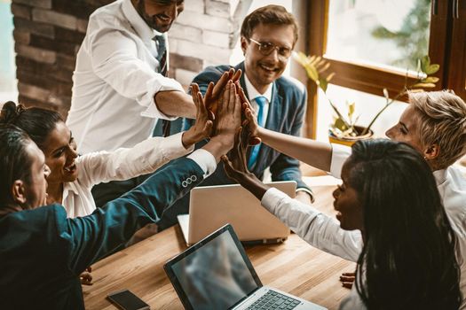 High Five Gesture Of Unity From Multi-Ethnic Business Team, High Angle View Of Cheerful Business People Joining Hands Together While Sitting At Office Table, Selective Focus on Human Hands In Centre