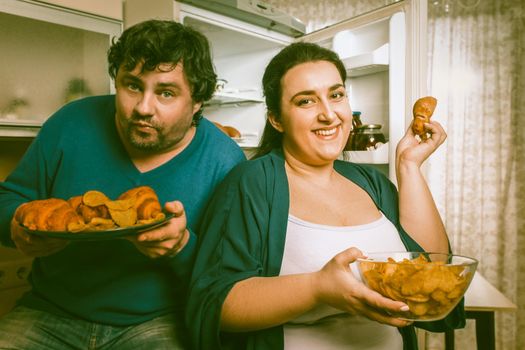 Body Positive Couple Having Fun With Unhealthy Eating In Kitchen, Smiling Man And Woman Standing Near Opened Refrigerator Holding Plate With Croissants And Bowl Of Potato Chips While looking At Camera