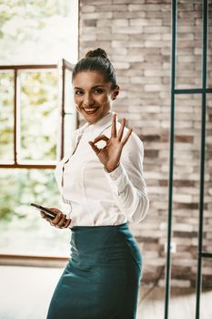 Young Business Woman Shows Ok And Toothy Smiles Looking At Camera, Portrait Of Cheerful Well-dressed Woman Holdind Smart Phone While Standing Near Window In Office Indoors, Toned Image