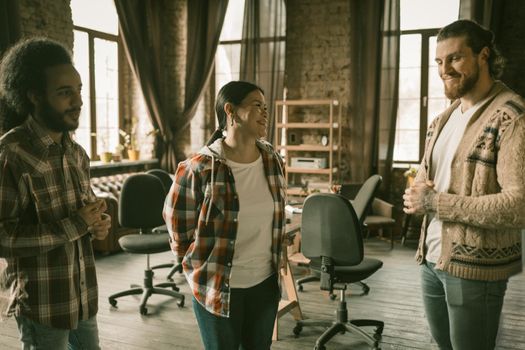 Cheerful Group Of Freelancers Or Students Standing In Coworking Space, Caucasian Young Man, Asian Woman And Arab Guy Have Fun Talking To Each Other In Loft Style Interior