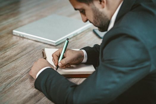 Businessman Writes In Note Pad Sitting At Desk, Close Up Shot Of Business Man Holding Pen And Doing Paperwork In His Office, Side View Of Office Worker, Toned Image