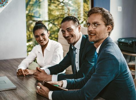 Team Of Office Worker Looks At Camera Sitting At Office Desk, Selective Focus On Smiling Businessman In Eyeglasses In Foreground, Woman And Two Men Brainstorming On Startup Strategy, Toned Image