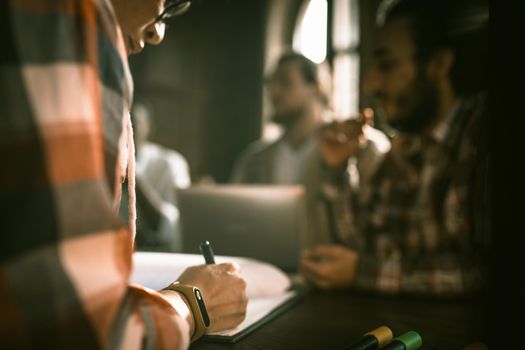 Brainstorming Of Diverse Freelancers Team Sitting At Table In Board Room, Abstract Defocused Image Of Unrecognizable Person With Female Hands Writing On Paper In Foreground,