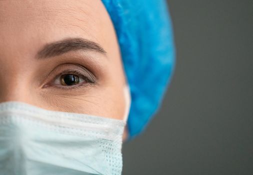 Female medical worker In Protective Mask And Blue Workwear Looking At Camera, Close Up Shot Of Cheerful woman's Eye, Copy Space Are At Right Side On Grey Back