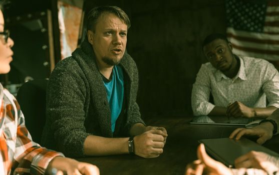 Caucasian Speaker Presents His Concept Of New Business, His Colleagues African Man And Asian Woman Listening To Him Carefully Sitting At Wooden Table With Digital Tablet On It, Focus On Speaker