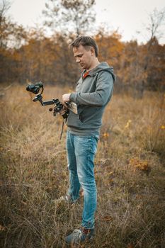 Film Maker Prepares His Camera On Digital Stabilizer For Shooting Video Outdoors, Caucasian Cameraman Or Blogger Prepare Equipment For Shooting Video Content On Blog Standing On Grass In Nature