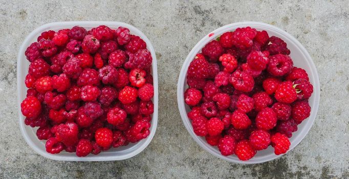 Square and round plastic transparent containers with raspberries on a dirty concrete background.