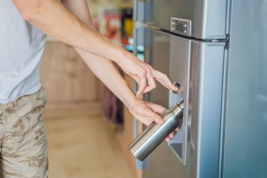 Male hand is pouring cold water and ice cubes in a metal bottle from dispenser of home fridge.