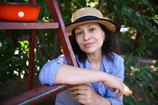 Portrait of a serene beautiful woman, farmer in straw hat, leaning on a ladder in a cherry orchard and looking at camera. Harvesting ripe cherries in an organic garden. Eco farming. Country lifestyle