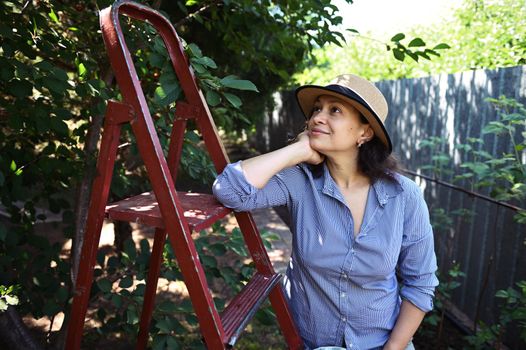 Pleasant multi-ethnic young woman sitting on the ladder and looking away dreamily while relaxing after picking cherries in the organic fruit garden. Harvest time. Agriculture. Horticulture. Gardening.