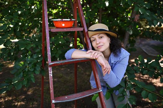 Charming Caucasian woman gardener, farmer in straw hat, leaning on a ladder in a cherry orchard and looking at camera. Harvesting ripe sweet cherries in an organic eco farm for sale in farmers markets