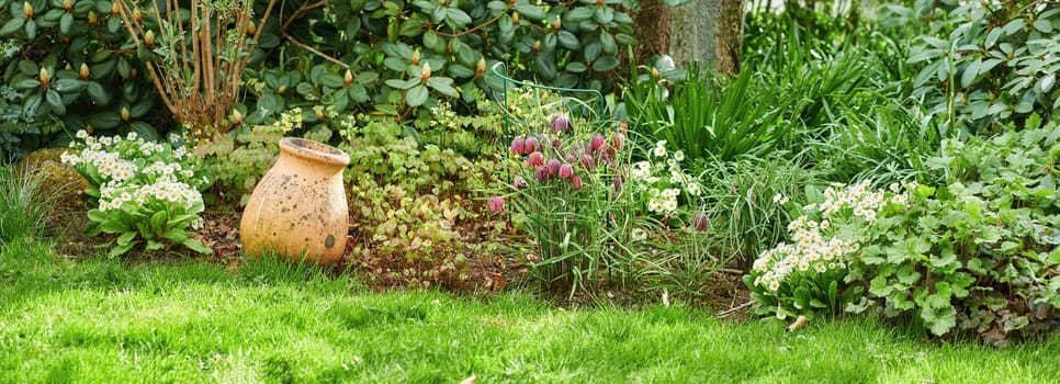 Green garden with a lush green meadow and flowering plants. Empty brown clay vase outdoors in a backyard or park on a summer day. Aged container outside in a bright vibrant bush or shrub in nature.