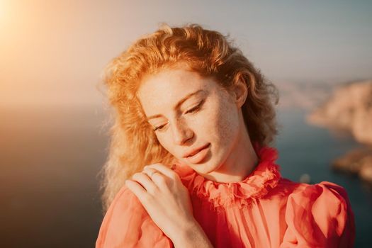 Close up shot of beautiful young caucasian woman with curly blond hair and freckles looking at camera and smiling. Cute woman portrait in a pink long dress posing on a volcanic rock high above the sea