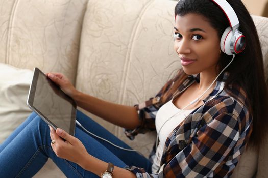 A dark-skinned girl in headphones with a tablet in her hands sits on the couch, close-up. Homeschooling during a pandemic